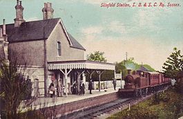 A vintage railway station with a small building, a covered platform, and a steam locomotive on the tracks.
