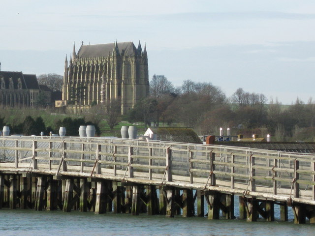 A wooden pier extends over calm water with a Gothic-style stone church perched on a hill in the background.