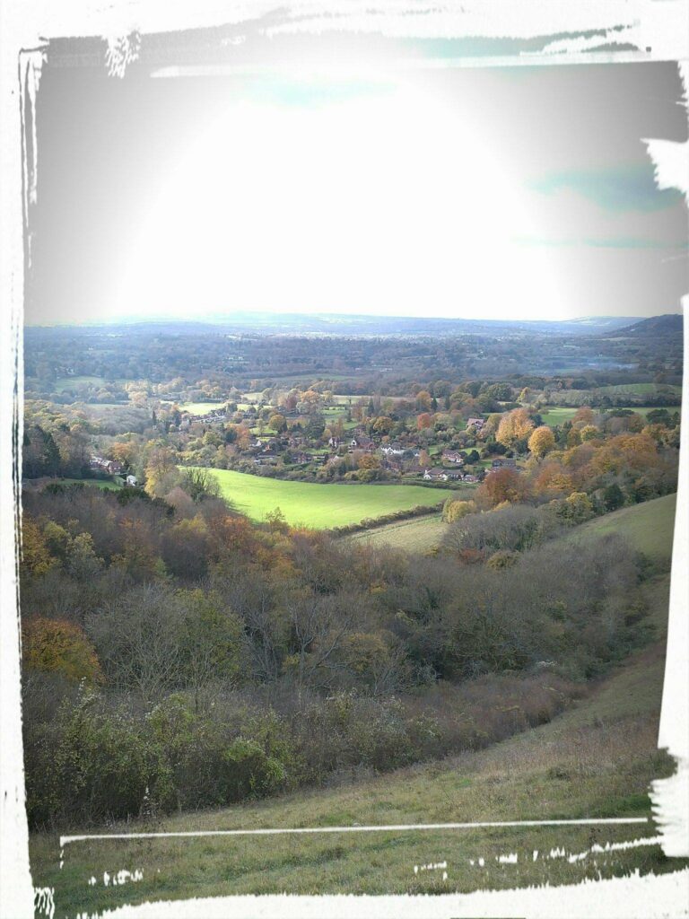 Sunlit countryside landscape with rolling hills, green fields, scattered trees, and a distant village on the horizon.