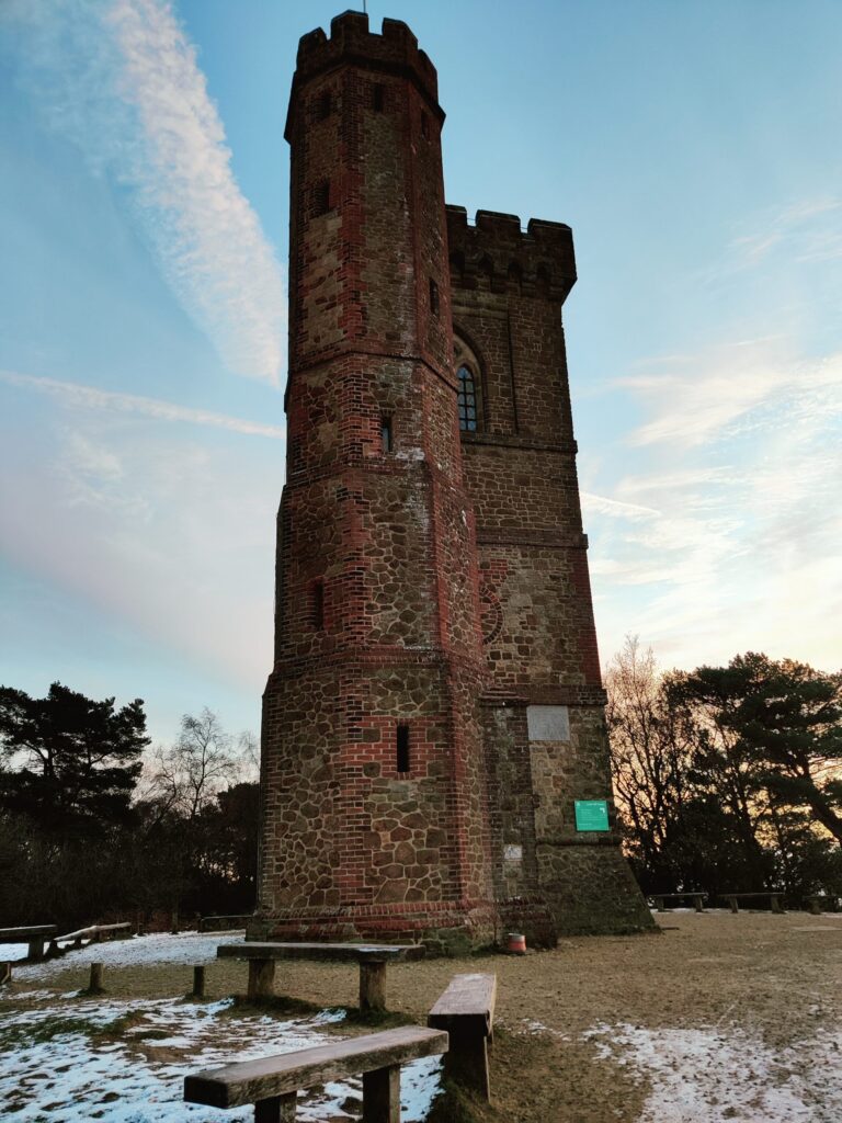 A tall brick tower with battlements stands in a snowy park under a blue sky with trees in the background.