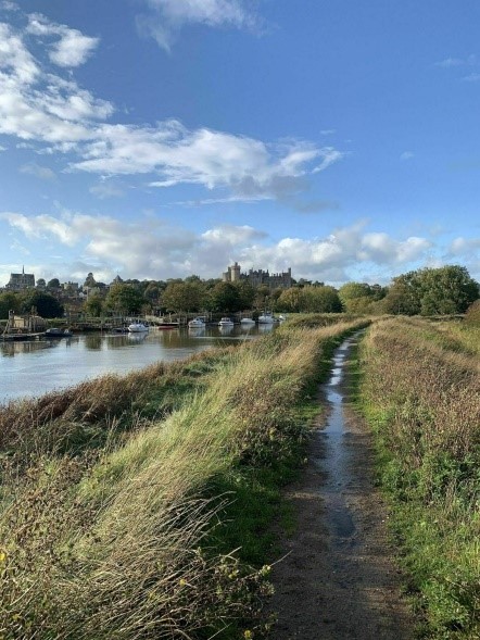 A narrow dirt path runs along a grassy canal with boats on the left and tall grasses on the right under a blue, partly cloudy sky.